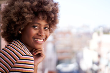 Young African American woman looking smiling on camera. Cheerful black afro girl leaning on home window. Portrait female beautiful expression posing for photo. Copy space and blurred background.
