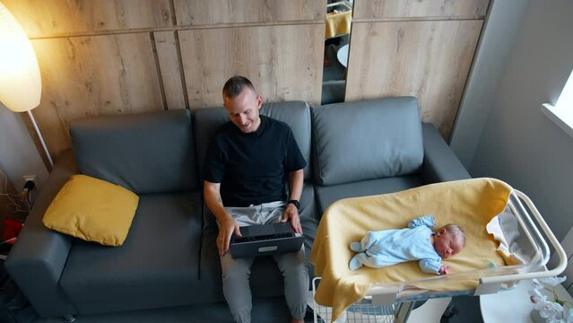 New-made Father Sitting On The Sofa Opens His Laptop. Tiny Newborn Child Lies In The Crib Beside. Top View.