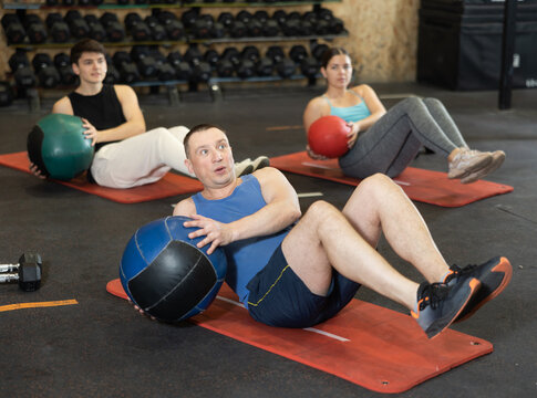Focused adult man taking part in high-intensity group training session, doing V-sit exercise with medicine ball and twisting torso to strengthen abdominal and oblique muscles..