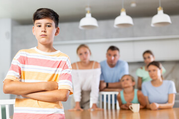 Fototapeta premium Sullen boy child crossed arms crossed over chest and is standing with back to parents and sisters. Large family is outraged by negligent behavior of teenage son