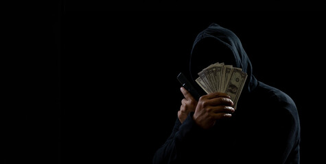 Portrait man thief wearing a black hood shirt, standing hand holding gun and money cash, counting the amount obtained from hijacking or robbing, in pitch-black room. dark background - obrazy, fototapety, plakaty