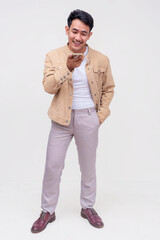 A handsome asian dude in a khaki jacket, white shirt and light gray pants. Holding a cellphone talking to someone via speaker phone. Whole body photo, isolated on a white background.