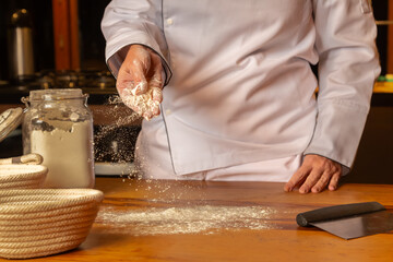 Chef de cozinha usando uma dolmã branco, preparando a bancada de madeira , jogando farinha para abrir a massa do pão, com detalhe na farinha parada no ar.