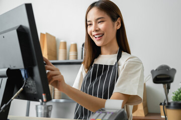Happy young Asian woman cashier wears an apron and using pos terminal to input orders on coffee...