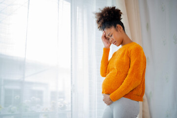 African American pregnancy woman standing beside glass window, hands at head and belly, eyebow fold...