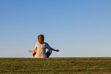 Woman sitting in the lotus pose on the green grass over blue sky, connecting with nature