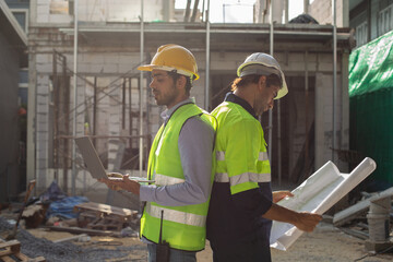 Construction engineer team. Two man engineer wear uniform holding blueprint and laptop standing at construction site. Workers supervise construction inspection of building.