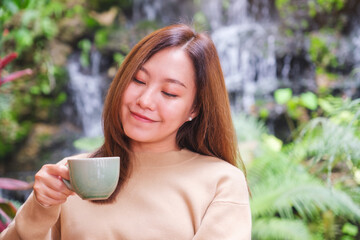 Portrait image of a young woman holding and drinking coffee while sitting in the garden with waterfall