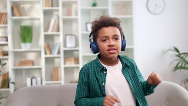 Active boy of multiracial ethnicity enjoying music via wireless headphones and briskly dancing in bright spacious room. Energetic child having fun at home. Concept of kids and modern technology