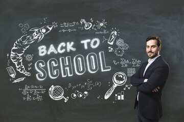 Attractive young businessman with folded arms standing on creative back to school sketch on blackboard background. Education, knowledge, and wisdom concept.