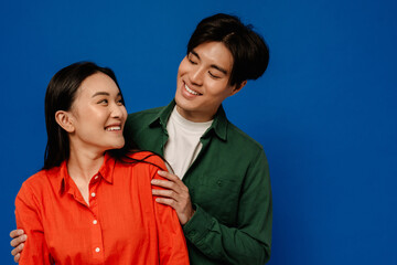 Obraz premium Smiling asian couple hugging while standing isolated over blue background