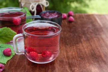 Hot raspberry tea in two transparent cups on a wooden table with blurred green background. Fresh berries, leaves and jars of jam. Side view, place for text copy space.