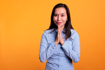 Positive asian woman praying to god in front of camera, holding hands in prayer gesture and asking for good luck. Religious model expressing hope and belief, standing in studio over yellow background