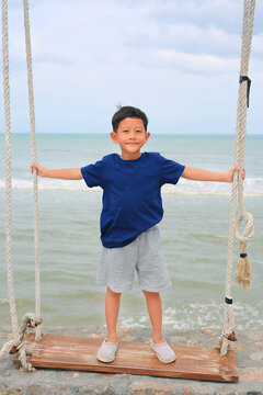 Asian little boy play standing on swing against cement wall at seaside.