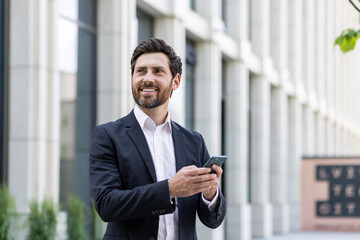 Young cheerful male businessman stands outside office buildings and uses the phone, reads news,...
