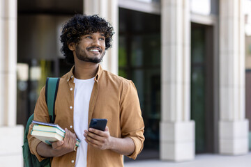 Smiling young Indian male student standing outside campus, holding books, backpack and using phone. Looks to the side