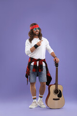 Stylish hippie man with guitar pointing at something on violet background