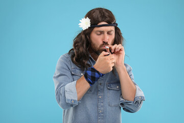 Stylish hippie man lighting cigarette on light blue background