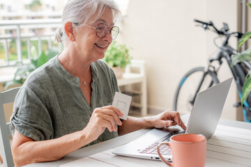 Online shopping concept. Happy senior woman using laptop sitting outdoors on terrace doing e-commerce shopping, senior woman in glasses holding credit card using modern technology