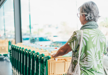 Retired senior woman in green color taking a shopping cart in supermarket for grocery purchases. Consumerism, inflation, rising costs concept