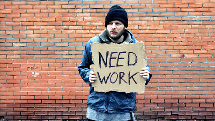 Homeless holds piece of cardboard with inscription Need Work. Refugee is at brick wall looking for...