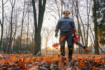 Mature man in blue uniform operating portable leaf blower outdoors. Bottom view of gray haired...
