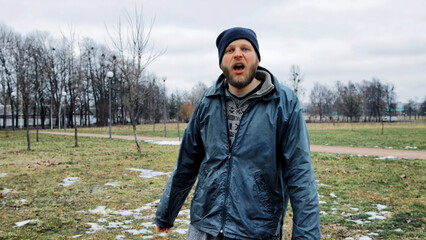 Angry drunk homeless man cursing, shouting and waving his hands in city park. Drunkenness,...
