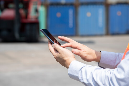 Technology concept, A man, shipping worker touching smart phone screen to manage an online order at the container dock, stock, freight and cargo supply chain logistics