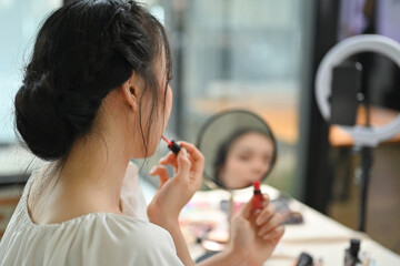 Young woman applying lipstick on lips in front of a mirror and recording make up tutorial with...
