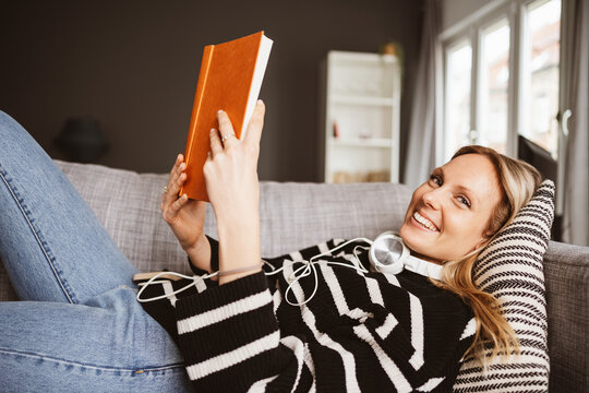 Laughing Young Woman with Headphones Lying on the Sofa and Reading a Book