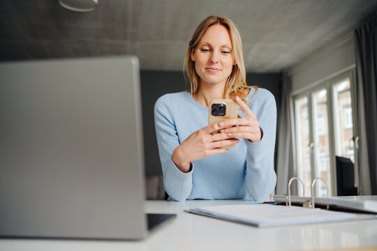 Blonde Young Woman at Office Desk, Looking at Her Smartphone