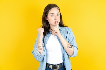 Young beautiful woman wearing denim overshirt asking to be quiet with finger on lips pointing with...