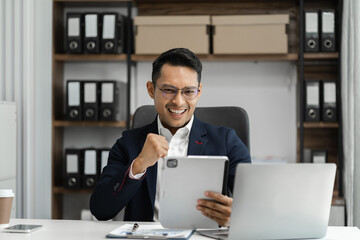 Fototapeta na wymiar Happy young businessman looking at successful excited tablet sitting at desk raising hands in yes gesture celebrating business success.