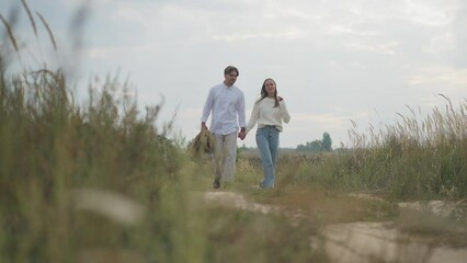 Wide shot young man strolling with woman covering spouse with jacket in slow motion leaving with partner. Happy loving confident Caucasian couple walking in autumn field outdoors