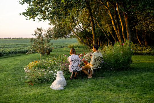 Man and woman have a romantic picnic on a beautiful green lawn with flowers and their dog sitting nearby. Spending summer time together on nature