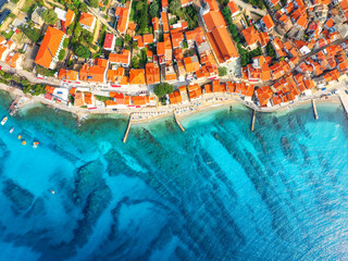 Aerial view of buildings with orange roofs, beach, clear blue sea in summer day. Vacation in beautiful Baska city, Krk island, Croatia. Top drone view of houses, streets, promenade, adriatic sea bay