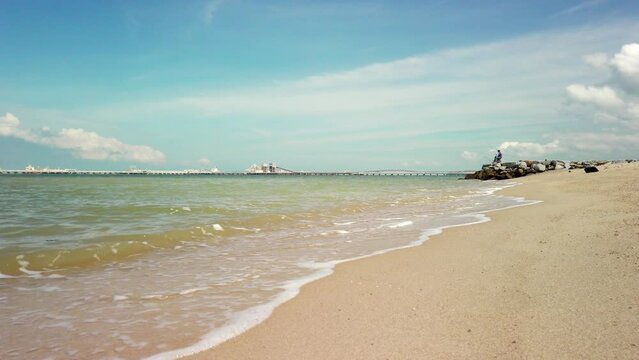 Daytime sea view at Pantai Puteri Beach, Melaka, Malaysia. Unidentified person fishing on rocks, with a refinery in the background.