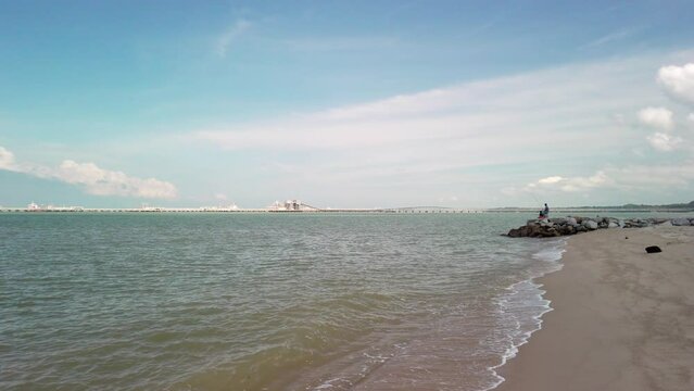 A drone-like low-to-high angle shot captures the coastal serenity of Pantai Puteri Beach in Melaka, Malaysia, during the daytime. In the background, a refinery adds industrial backdrop to the scene.