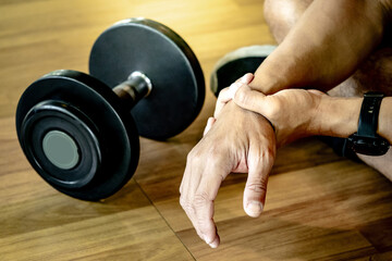 Male athlete massaging his wrist and hand suffering from wrist pain caused by sprain or joint fracture from overweight dumbbell lifting in fitness gym. Sport injury concept - obrazy, fototapety, plakaty