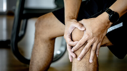 Sportive man feeling knee pain or rheumatoid arthritis while sitting on workout bench in fitness...