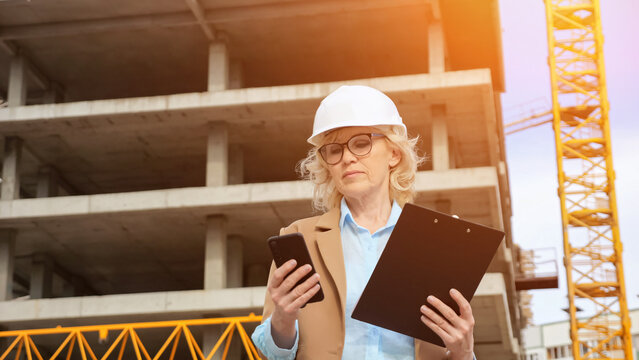 Senior woman entrepreneur in protective helmet types data to client on phone from clipboard on background of building. Concept of construction and working moments, sunlight