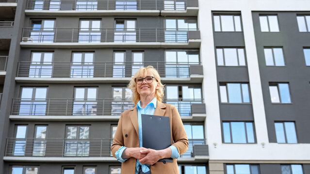 Mature businesswoman stands holding clipboard in hands against residential complex in daytime. Female real estate agent posing for camera low angle shot