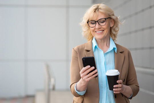 Smiling senior businesswoman with cup of coffee in formal dress responds to message on smartphone. Blonde female person works during coffee break, copyspace