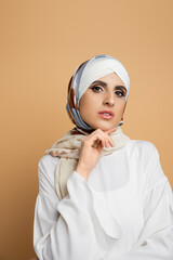 sensual muslim woman in stylish outfit posing with hand near chin and looking at camera on beige