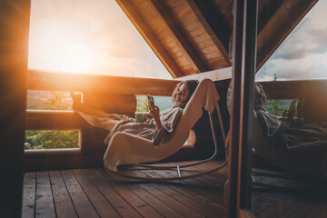 Young relaxed cheerful woman enjoying nature on the balcony of the cabin at sunset with a cup of tea and a smartphone in her hands