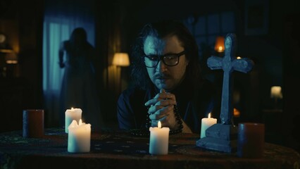 Shot capturing a table with magical tools on it. A priest is sitting at the table with beads in his hands, praying with his eyes closed. Female silhouette, ghost approaches him.