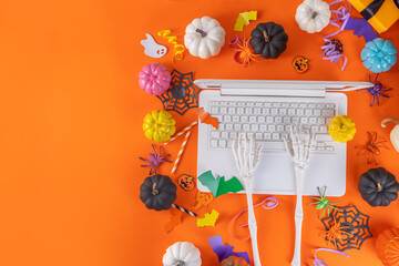 Funny Halloween bright yellow composition. White laptop with typing skeleton hands, Halloween decor colorful holiday accessories - spiders, cobwebs, pumpkins, bats, top view copy space banner
