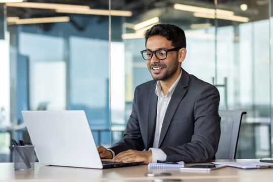Young successful Arab businessman in business clothes works inside the office with a laptop, satisfied with the results, the boss in glasses types on the computer keyboard