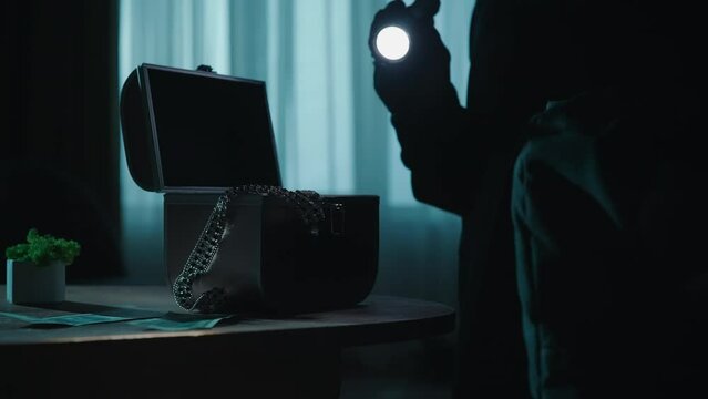 A thief commits a robbery in a dark apartment, using a flashlight. Hands of a criminal stealing money and jewelry from a jewelry box close up. An alarm goes off, a blue ed light flashes.