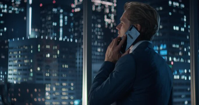 Backview of Handsome Caucasian Businessman in Stylish Suit Standing Next to Window in Big City Office with Skyscrapers Late At Night. Male CEO Talking On Smartphone With Partners.
